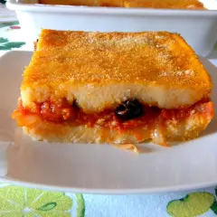 Una fetta di pitta di patate dorata e ripiena di sugo, cipolle e un'oliva nera, servita su un piatto bianco.