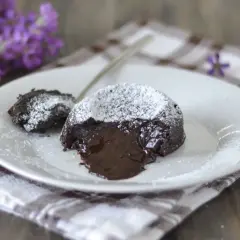 Un delizioso tortino al cioccolato con cuore fuso, spolverato di zucchero a velo, su un piatto bianco con fiori viola sullo sfondo.