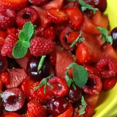 Macedonia di fragole, lamponi e ciliegie, guarnita con foglie di menta fresca, in una ciotola gialla.