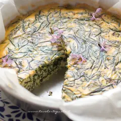 Frittata di agretti appena sfornata, decorata con fiori viola, in una teglia foderata di carta forno.
