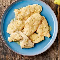 Piatto di petto di pollo al limone, condito con erbe aromatiche e servito su un piatto blu. Accanto, limone fresco e pepe nero.