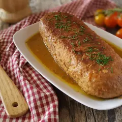 Polpettone dorato in padella, guarnito con erba cipollina, su un piatto bianco con salsa, accanto a pomodorini e una spatola di legno.