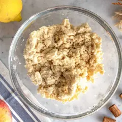 Impasto friabile per crumble di mele in una ciotola di vetro, con limone, mela e cannella sullo sfondo.