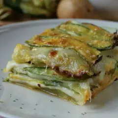 Una fetta di parmigiana di zucchine bianca, stratificata con formaggio filante e zucchine grigliate, servita su un piatto bianco.