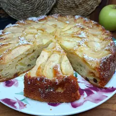 Una fetta di torta di mele, cosparsa di zucchero a velo, su un piatto fiorato. Sullo sfondo, una mela verde.