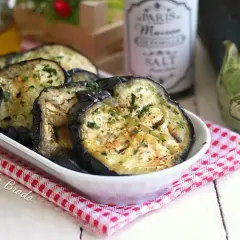 Fette di melanzane in padella, dorate e profumate con prezzemolo, servite in una ciotola bianca su una tovaglietta a quadretti rossi.