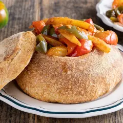 Un pane scavato ripieno di un saporito stufato di pipi e patate, con una fetta di pane accanto.