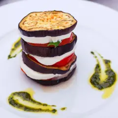 Parmigiana di melanzane grigliate a strati con mozzarella e pomodoro, guarnita con pesto e olio.