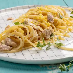 Un piatto di pasta tonno e limone, condita con scorza di limone grattugiata e erbe aromatiche fresche, servita su un piatto a scacchi bianchi.