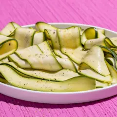 Fettine sottili di zucchine marinate crude, condite con pepe nero, disposte a spirale in un piatto bianco su un tavolo rosa acceso.