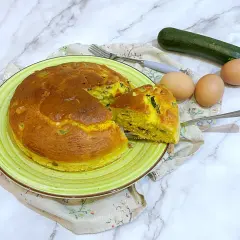Torta salata zucchine e speck dorata su piatto verde, con ingredienti freschi a lato.