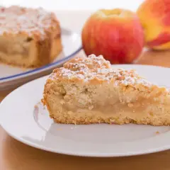 Una fetta di sbriciolata di mele, cosparsa di zucchero a velo, su un piatto bianco. Sullo sfondo, una torta intera e mele rosse.