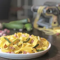 Piatto di pasta fresca con zucchine e pancetta, ingredienti freschi e macchina per la pasta sullo sfondo.