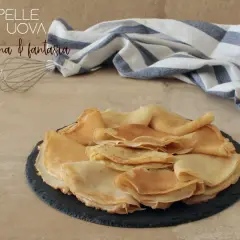 Crepes senza uova, dorate e soffici, impilate su un piatto scuro. Un battitore e un tovagliolo a righe completano la scena.