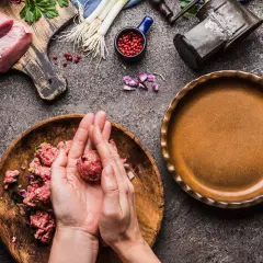 Mani che formano polpette di carne su un piatto di legno, con ingredienti freschi e una padella pronta.