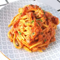 Un piatto di spaghetti al tonno, conditi con una ricca salsa rossa e guarniti con prezzemolo tritato, serviti su un piatto con motivo a squame.
