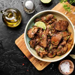 Piatto di fegatini di pollo saltati con cipolle, guarniti con prezzemolo fresco e spicchi di lime. Accanto, olio d'oliva, sale grosso e pepe.