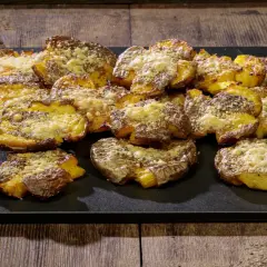 Patate schiacciate al forno, dorate e croccanti, cosparse di formaggio grattugiato e erbe aromatiche, servite su una pietra scura.