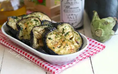 Fette di melanzane in padella, dorate e profumate con prezzemolo, servite in una ciotola bianca su una tovaglietta a quadretti rossi.