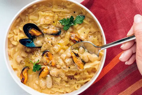 Un cucchiaio raccoglie una porzione di pasta fagioli e cozze, un piatto ricco e saporito, guarnito con prezzemolo fresco.