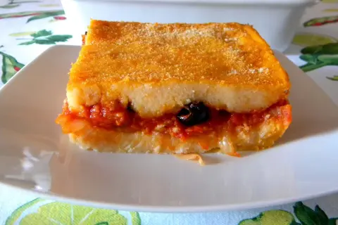 Una fetta di pitta di patate dorata e ripiena di sugo, cipolle e un'oliva nera, servita su un piatto bianco.