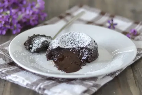 Un delizioso tortino al cioccolato con cuore fuso, spolverato di zucchero a velo, su un piatto bianco con fiori viola sullo sfondo.