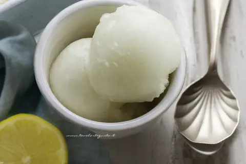 Coppetta con due palline di sorbetto al limone senza albume, accanto una fetta di limone e un cucchiaio decorato.