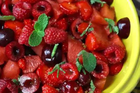 Macedonia di fragole, lamponi e ciliegie, guarnita con foglie di menta fresca, in una ciotola gialla.