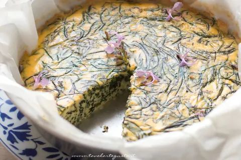 Frittata di agretti appena sfornata, decorata con fiori viola, in una teglia foderata di carta forno.