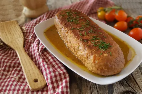 Polpettone dorato in padella, guarnito con erba cipollina, su un piatto bianco con salsa, accanto a pomodorini e una spatola di legno.