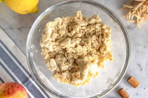 Impasto friabile per crumble di mele in una ciotola di vetro, con limone, mela e cannella sullo sfondo.