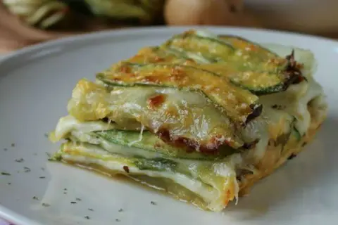Una fetta di parmigiana di zucchine bianca, stratificata con formaggio filante e zucchine grigliate, servita su un piatto bianco.