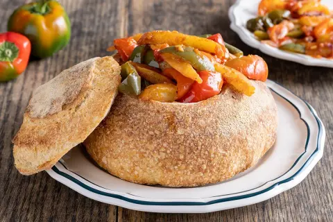 Un pane scavato ripieno di un saporito stufato di pipi e patate, con una fetta di pane accanto.