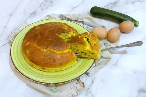 Torta salata zucchine e speck dorata su piatto verde, con ingredienti freschi a lato.