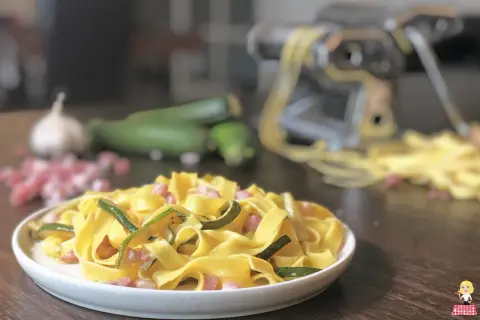 Piatto di pasta fresca con zucchine e pancetta, ingredienti freschi e macchina per la pasta sullo sfondo.