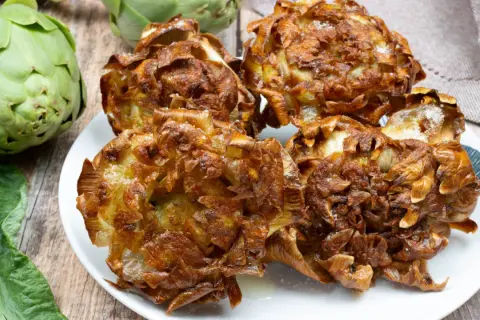 Carciofi alla giudia dorati e croccanti su un piatto bianco, con carciofi freschi sullo sfondo.