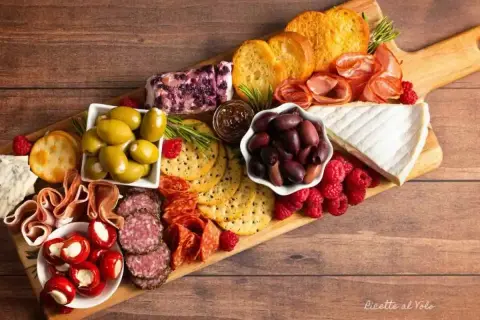 Un invitante tagliere salumi e formaggi con olive, lamponi, pane tostato e peperoncini ripieni.