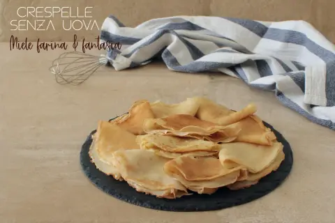Crepes senza uova, dorate e soffici, impilate su un piatto scuro. Un battitore e un tovagliolo a righe completano la scena.