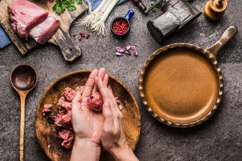 Mani che formano polpette di carne su un piatto di legno, con ingredienti freschi e una padella pronta.