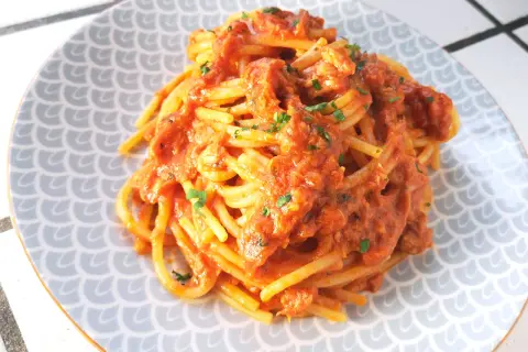 Un piatto di spaghetti al tonno, conditi con una ricca salsa rossa e guarniti con prezzemolo tritato, serviti su un piatto con motivo a squame.