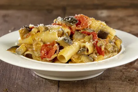 Piatto di pasta con melanzane e pomodorini, condita con formaggio grattugiato, su un tavolo di legno.