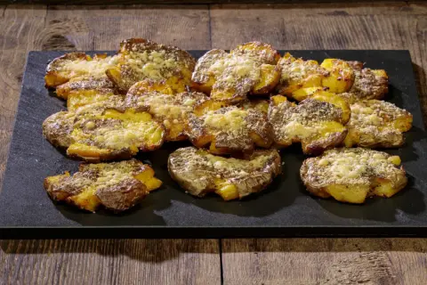 Patate schiacciate al forno, dorate e croccanti, cosparse di formaggio grattugiato e erbe aromatiche, servite su una pietra scura.