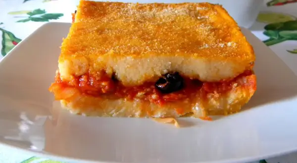 Una fetta di pitta di patate dorata e ripiena di sugo, cipolle e un'oliva nera, servita su un piatto bianco.