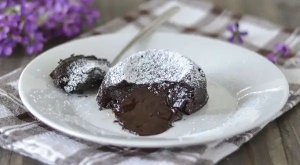 Un delizioso tortino al cioccolato con cuore fuso, spolverato di zucchero a velo, su un piatto bianco con fiori viola sullo sfondo.