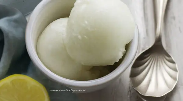Coppetta con due palline di sorbetto al limone senza albume, accanto una fetta di limone e un cucchiaio decorato.