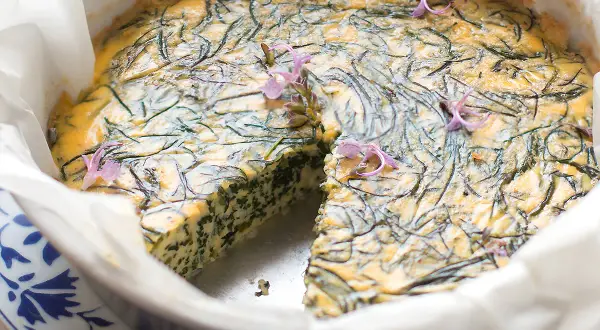 Frittata di agretti appena sfornata, decorata con fiori viola, in una teglia foderata di carta forno.