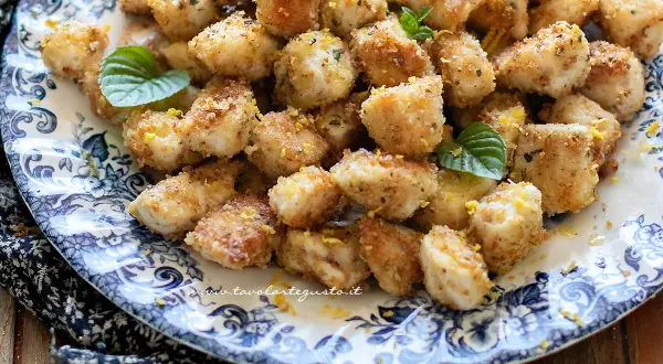 Bocconcini di pollo al forno dorati e croccanti, guarniti con scorza di limone e foglie di menta fresca su un piatto decorato.