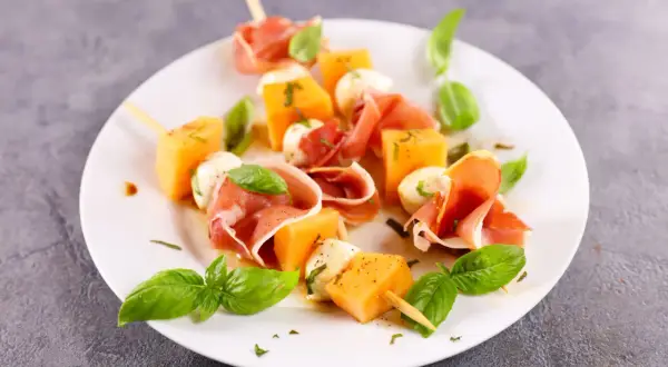 Spiedini di prosciutto e melone con mozzarelline e basilico fresco, un antipasto estivo perfetto.