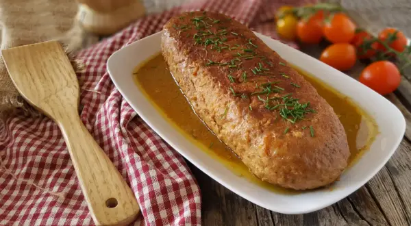 Polpettone dorato in padella, guarnito con erba cipollina, su un piatto bianco con salsa, accanto a pomodorini e una spatola di legno.