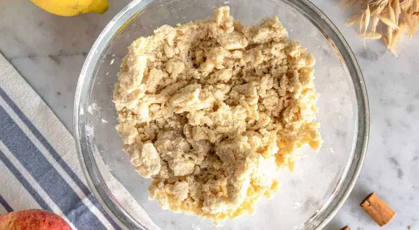 Impasto friabile per crumble di mele in una ciotola di vetro, con limone, mela e cannella sullo sfondo.